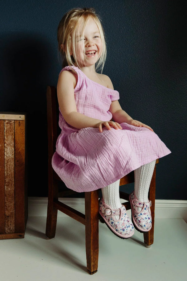 Young toddler in a pink linen dress sitting on a wooden chair against a dark wall.