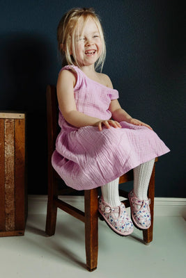 Young toddler in a pink linen dress sitting on a wooden chair against a dark wall.