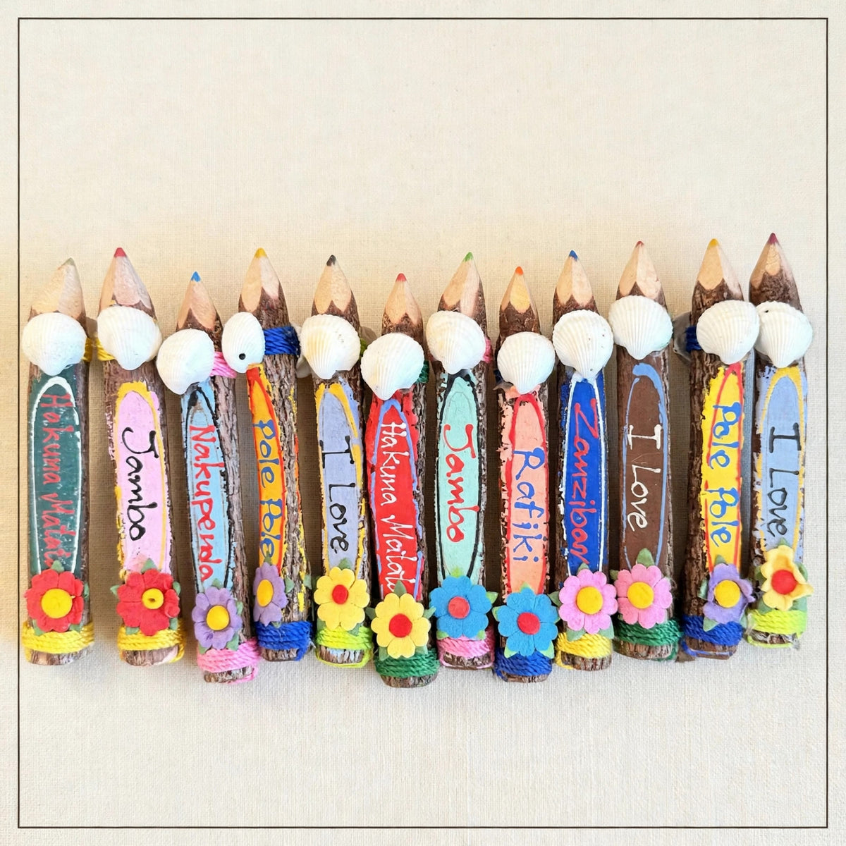 Row of small hand-carved wooden coloring pencils from Zanzibar on cream linen, hand-painted Swahili words, real seashells and felt flowers